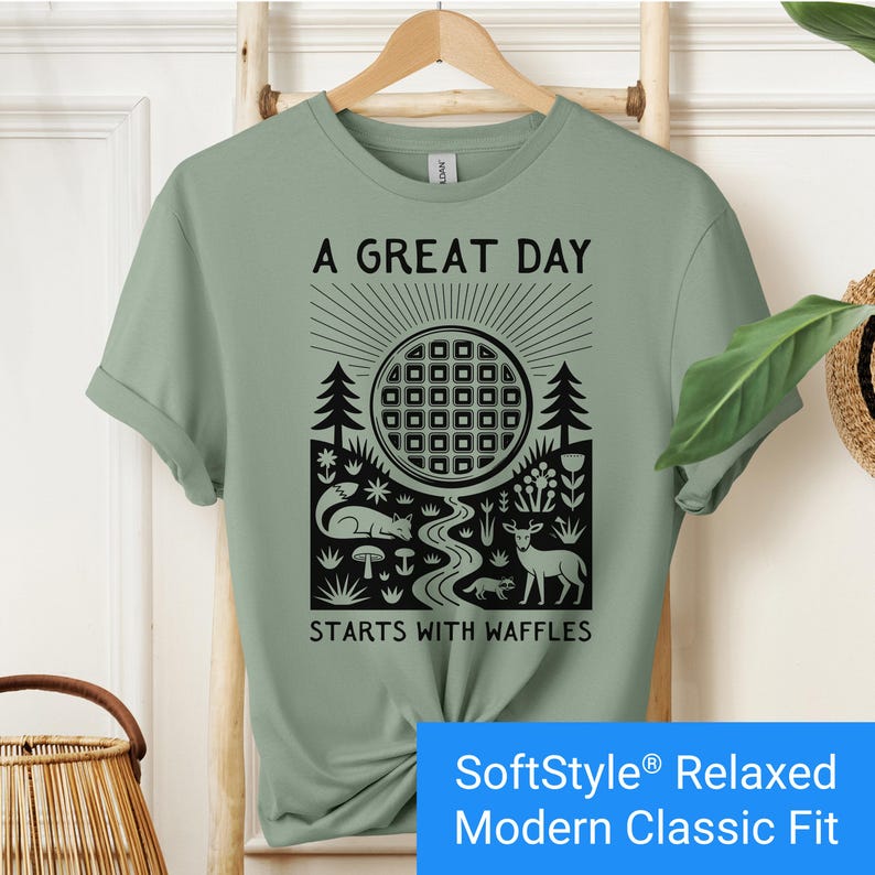 Sage color t-shirt featuring a folk art style graphic of a round waffle rising like a sun above a forest scene with woodland animals, plants, and a stream. Text above reads “A GREAT DAY” and below reads “STARTS WITH WAFFLES.”