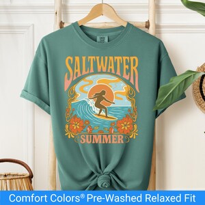 Light green t-shirt featuring a retro-style design of a female surfer riding a wave in front of an orange sunset, surrounded by floral and ocean motifs with the words "Saltwater Summer" in bold vintage fonts.