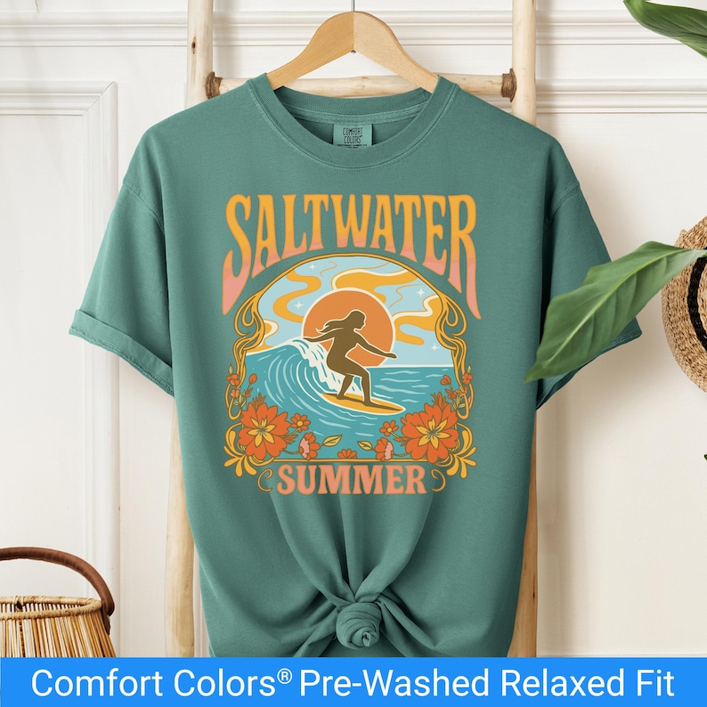 Light green t-shirt featuring a retro-style design of a female surfer riding a wave in front of an orange sunset, surrounded by floral and ocean motifs with the words "Saltwater Summer" in bold vintage fonts.