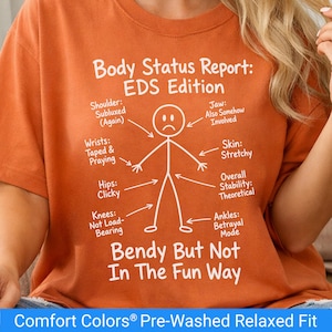 Op de afbeelding: Oranje t-shirt met witte tekst en een stokfiguurdiagram. De tekst luidt "Body Status Report: EDS Edition" met verschillende lichaamsdelen gelabeld. Daaronder staat "Bendy But Not In The Fun Way."