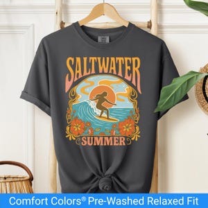 Pepper color t-shirt featuring a retro-style design of a female surfer riding a wave in front of an orange sunset, surrounded by floral and ocean motifs with the words "Saltwater Summer" in bold vintage fonts.