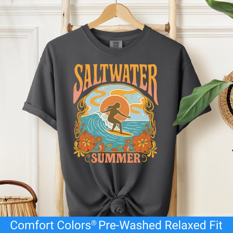 Pepper color t-shirt featuring a retro-style design of a female surfer riding a wave in front of an orange sunset, surrounded by floral and ocean motifs with the words "Saltwater Summer" in bold vintage fonts.