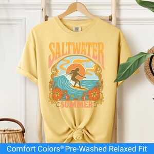 Butter color t-shirt featuring a retro-style design of a female surfer riding a wave in front of an orange sunset, surrounded by floral and ocean motifs with the words "Saltwater Summer" in bold vintage fonts.