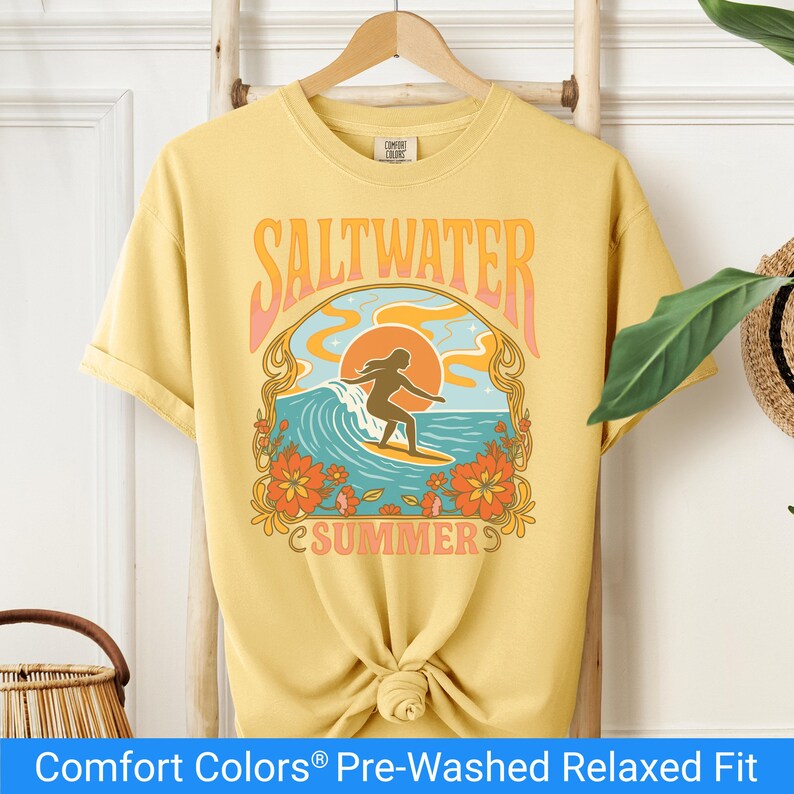 Butter color t-shirt featuring a retro-style design of a female surfer riding a wave in front of an orange sunset, surrounded by floral and ocean motifs with the words "Saltwater Summer" in bold vintage fonts.