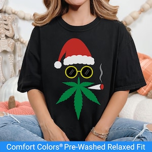May include: Black t-shirt featuring a Christmas-themed design. The graphic includes a red Santa hat, yellow-rimmed glasses, a green cannabis leaf, and a lit joint. The text "Comfort Colors® Pre-Washed Relaxed Fit" is at the bottom.