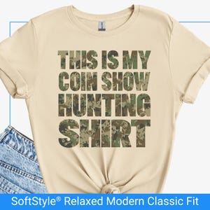 Könnte beinhalten: Hellbeige T-Shirt mit dem Text "THIS IS MY COIN SHOW HUNTING SHIRT" in einem Camouflage-Muster. Das Shirt hat kurze Ärmel und eine lockere Passform.