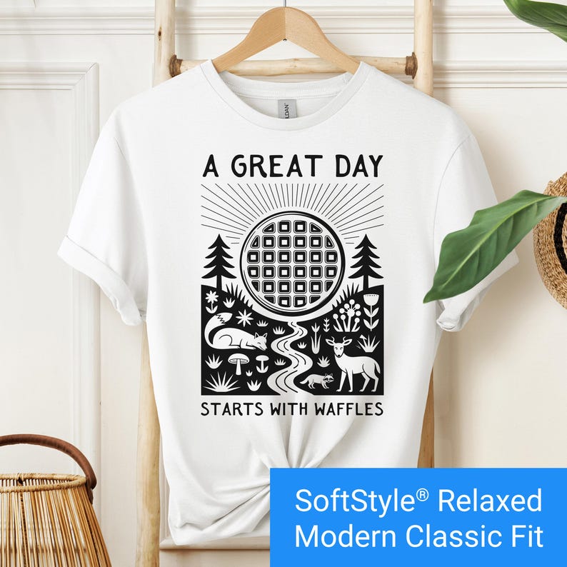 White t-shirt featuring a folk art style graphic of a round waffle rising like a sun above a forest scene with woodland animals, plants, and a stream. Text above reads “A GREAT DAY” and below reads “STARTS WITH WAFFLES.”
