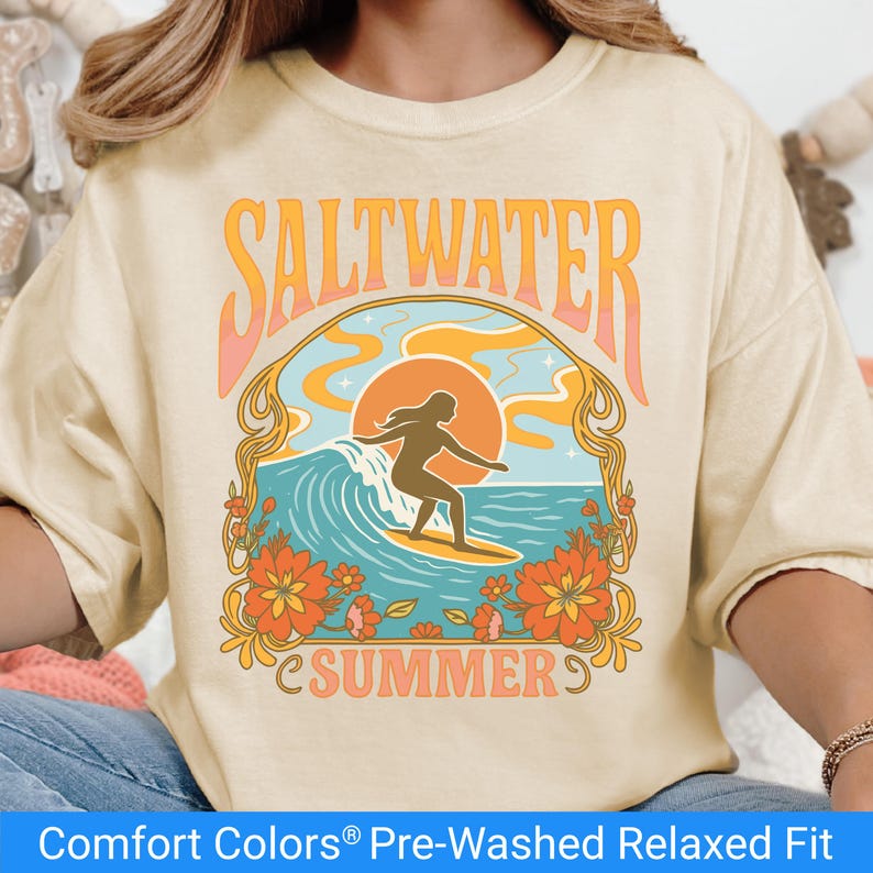 Ivory color t-shirt featuring a retro-style design of a female surfer riding a wave in front of an orange sunset, surrounded by floral and ocean motifs with the words "Saltwater Summer" in bold vintage fonts.