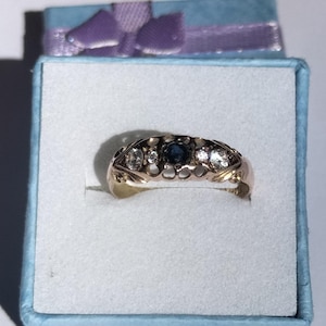 Op de afbeelding: Een goudkleurige ring met een centrale donkerblauwe edelsteen, geflankeerd door kleinere, heldere stenen. De ring is gezet in een decoratieve band met opengewerkte details. Hij ligt in een lichtblauwe geschenkdoos met een paars lint.