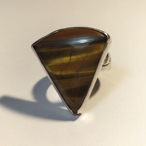 May include: A silver ring with a triangular tiger's eye gemstone. The stone has bands of brown, gold, and black. The ring has a simple, polished silver band. The gemstone is approximately 3.8 cm wide.