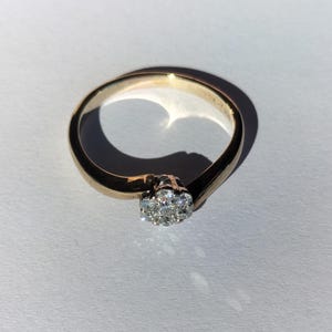 May include: A gold ring featuring a cluster of seven small, clear, round-cut stones set in a circular arrangement at the center. The band is smooth and polished, with a bypass-style design that tapers toward the central stone cluster setting.
