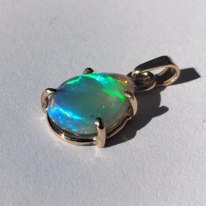 May include: A gold pendant with an oval opal gemstone. The opal exhibits a vibrant play of colour with shades of blue, green, and yellow. The gemstone is secured by four gold prongs, with a gold bail for a chain.