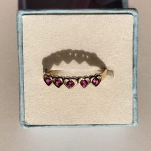 Puede incluir: Un anillo dorado con cinco gemas en forma de corazón de color rosa intenso. Las gemas están engastadas en marcos en forma de corazón. El anillo se presenta en una caja azul claro con interior blanco.