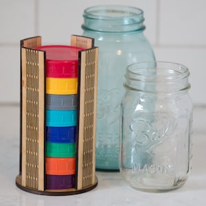 May include: A wooden storage organizer with a rainbow of colored lids for mason jars. The organizer has a grid pattern and is holding six lids in a rainbow color order: red, yellow, gray, blue, green, and purple.