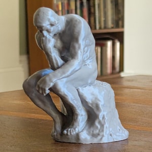 Op de afbeelding: Een lichtgrijze sculptuur van een zittende figuur, met de rechterhand die de kin aanraakt en de linkerarm rustend op het linkerbeen. De figuur zit op een rotsachtige basis. De sculptuur staat op een houten oppervlak.