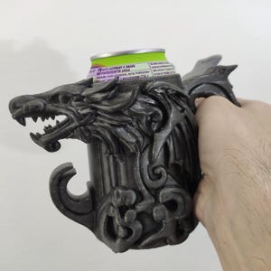 以下が含まれることがあります： オオカミの頭のデザインが施された、ダークグレーの3Dプリント缶ホルダー。ホルダーは、オオカミの牙をむいた顔と装飾的な模様が特徴で、細部までこだわっています。緑と白の缶がホルダーの中に置かれています。