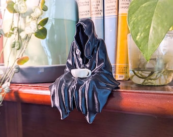 Puede incluir: Una figura negra de una figura con capa sosteniendo una pequeña taza de té blanca. La figura está sentada sobre una superficie de madera, con libros y plantas en el fondo. La figura tiene una capucha y ropa drapeada.