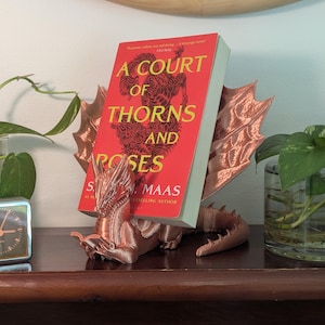 Könnte beinhalten: Ein kupferfarbener Drachen-Buchhalter mit einem Buch mit dem Titel "A Court of Thorns and Roses" im Halter. Das Buch hat einen roten Einband mit goldener Schrift. Der Drache steht auf einer Holzoberfläche mit Pflanzen und einer Uhr.