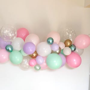 May include: A collection of pastel-colored balloons in pink, white, green, and purple, with some featuring star patterns. Metallic gold and teal balloons add accents. The balloons are arranged in a decorative cluster.
