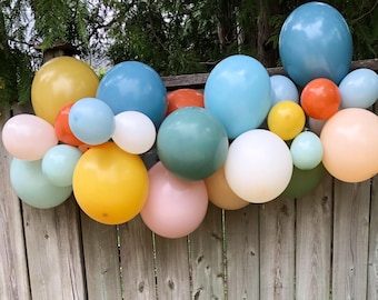 May include: A display of colourful balloons in different sizes and shades, including yellow, blue, orange, pink, and green. The balloons are arranged against a wooden fence, creating a festive atmosphere.