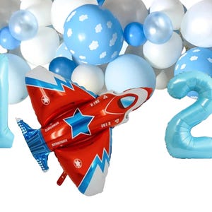 May include: A collection of blue and white balloons, including the numbers 1 and 2. A red and white airplane-shaped balloon with blue detailing and star designs is also featured. Ideal for a birthday party or celebration.