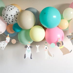 May include: A colourful balloon display featuring pastel shades of pink, green, and blue. A garland of dog-shaped cutouts, each wearing a party hat, is strung beneath the balloons, creating a celebratory scene.