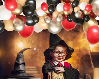 May include: A child wearing a black pointed hat, round glasses, and a black robe with a red tie, holding a book. Above the child is an arch of balloons in red, black, gold, and white. A black pointed hat sits on a surface to the left.