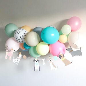 May include: A cluster of pastel-coloured balloons, including pink, blue, green, and yellow, with a paw print balloon. A garland of dog-shaped cutouts hangs below, creating a cheerful party decoration.