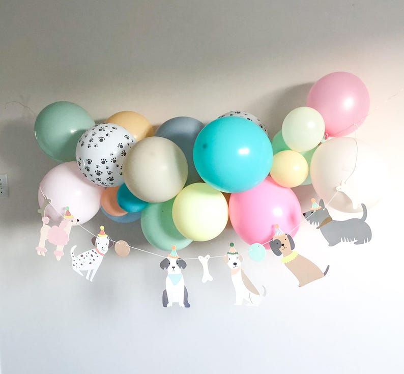 May include: A cluster of pastel-coloured balloons, including pink, blue, green, and yellow, with a paw print balloon. A garland of dog-shaped cutouts hangs below, creating a cheerful party decoration.