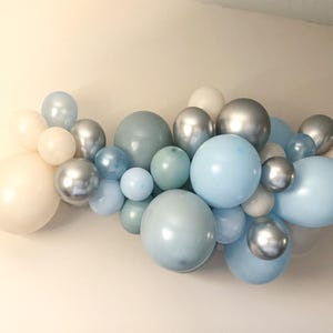 K&ouml;nnte beinhalten: Eine Ansammlung pastellfarbener Ballons in verschiedenen Gr&ouml;&szlig;en und Schattierungen von Blau, Silber und Creme. Die Ballons sind in einem Cluster angeordnet und bilden eine dekorative Darstellung. Mit matten und metallischen Oberfl&auml;chen.