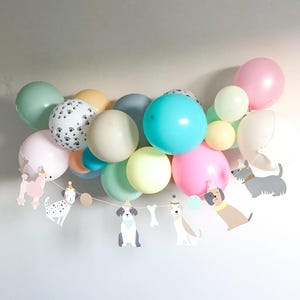May include: A cluster of pastel-coloured balloons, including pink, blue, green, and yellow, with a paw print balloon. A garland of dog-shaped cutouts wearing party hats is strung beneath the balloons, creating a celebratory scene.