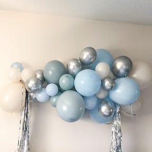K&ouml;nnte beinhalten: Eine Ansammlung von Ballons in verschiedenen Gr&ouml;&szlig;en und Farbt&ouml;nen von Blau, Silber und Wei&szlig;. Die Ballons sind in einer dekorativen Anordnung angeordnet, mit silbernen Fransen. Die Anordnung ist wahrscheinlich f&uuml;r eine Party oder Feier.