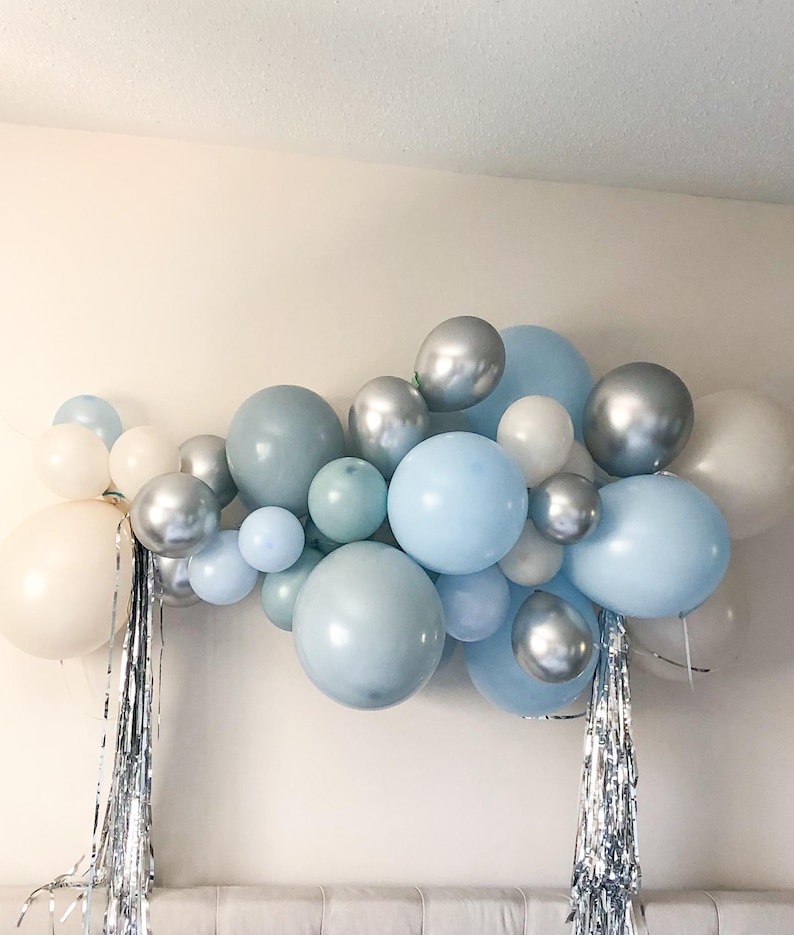 K&ouml;nnte beinhalten: Eine Ansammlung von Ballons in verschiedenen Gr&ouml;&szlig;en und Farbt&ouml;nen von Blau, Silber und Wei&szlig;. Die Ballons sind in einer dekorativen Anordnung angeordnet, mit silbernen Fransen. Die Anordnung ist wahrscheinlich f&uuml;r eine Party oder Feier.