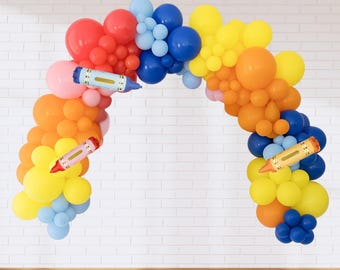May include: A vibrant balloon arch in shades of red, orange, yellow, blue, and pink, adorned with crayon-shaped balloons. Ideal for a party or classroom setting. The arch is a cheerful and eye-catching decoration.