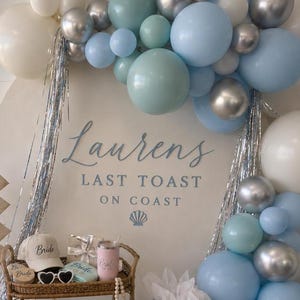 K&ouml;nnte beinhalten: Dekoration f&uuml;r eine Brautparty mit einem Ballonbogen in Blau, Silber und Wei&szlig;. Der Hintergrundtext lautet "Lauren's Last Toast on Coast". Zubeh&ouml;r: Hut, Sonnenbrille, Tragetaschen und Geschenke. Ma&szlig;e in cm.