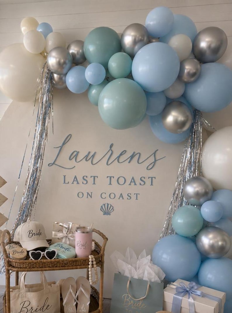 K&ouml;nnte beinhalten: Dekoration f&uuml;r eine Brautparty mit einem Ballonbogen in Blau, Silber und Wei&szlig;. Der Hintergrundtext lautet "Lauren's Last Toast on Coast". Zubeh&ouml;r: Hut, Sonnenbrille, Tragetaschen und Geschenke. Ma&szlig;e in cm.