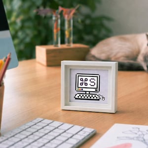 May include: A framed cross-stitch featuring a retro computer design with the Command key symbol. The artwork is in a white frame, placed on a wooden desk alongside a keyboard, pencils, and a sleeping cat. The color palette includes black, white, and various colors.