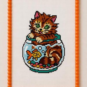 May include: A cross-stitch embroidery on white fabric featuring a brown tabby cat sitting inside a round glass fishbowl with a small yellow goldfish and colourful gravel. The piece is framed by a border of small, round, bright orange beads.
