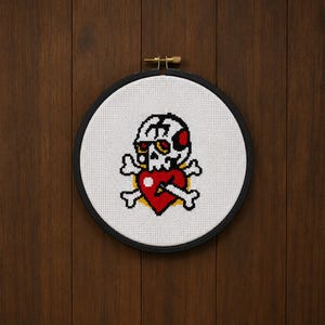 May include: A circular black embroidery hoop containing white Aida fabric with a cross-stitch design of a white skull wearing sunglasses, positioned above a red heart and two crossed white bones, all outlined in black thread on a white background.