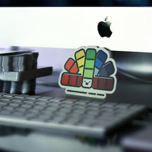 May include: A colorful sticker featuring a palette of paint swatches in red, orange, yellow, green, blue, and purple. The sticker has a smiling face and is placed on a keyboard in front of a computer monitor.