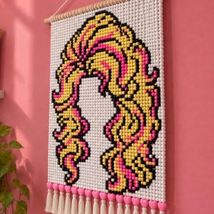 May include: A wall hanging made of white, yellow, pink, and black beads arranged to form a stylized graphic of long, wavy hair. The piece is suspended from a wooden dowel with a white string and features a row of pink heart beads and white tassels.