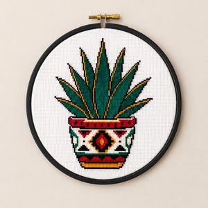 May include: A cross-stitch embroidery of a green agave plant in a decorative pot with red, white, black, and yellow geometric patterns, framed in a circular black hoop with a metal tension screw at the top, displayed on a white fabric background.