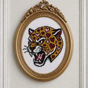 Puede incluir: Un bordado de punto de cruz de una cabeza de leopardo gruñendo con manchas en forma de corazón en tonos amarillo, rojo y rosa, sobre un fondo de tela blanca y enmarcado en un marco ovalado de color dorado con detalles florales.