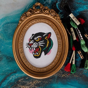 May include: An oval gold frame contains a cross-stitch of a panther's head. The panther is black with yellow eyes and a red mouth. Embroidery threads in black, green, and red are next to the frame. The background is teal and gold.