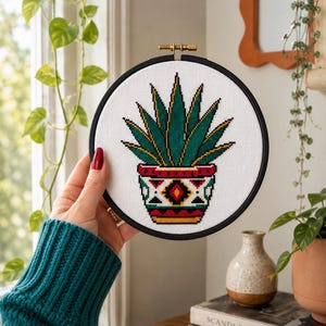 Peut inclure: Un cercle de broderie au point de croix représentant une plante succulente verte et jaune dans un pot décoratif. Le pot présente un motif géométrique rouge, noir et jaune. La broderie est dans un cercle noir avec un fermoir doré.