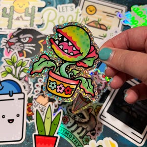 May include: A holographic sticker featuring a green carnivorous plant with an open mouth and sharp teeth, potted in a yellow container decorated with colorful flower icons. The sticker has a shimmering, glittery finish and bold black outlines.