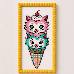 May include: A cross-stitch embroidery in a yellow beaded frame featuring a stacked ice cream cone design. The top scoop is a pink cat face with a cherry on top, and the bottom scoop is a teal cat face. The cone has a pink and yellow grid pattern.