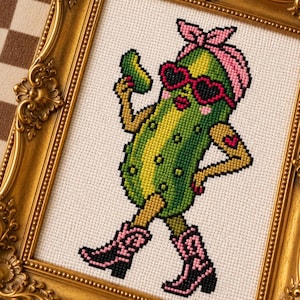 May include: A cross-stitch embroidery of a green pickle character wearing a pink bandana, heart-shaped sunglasses, and pink cowboy boots. The pickle is holding a small green object and has a heart tattoo on its arm. It is displayed in an ornate gold frame.