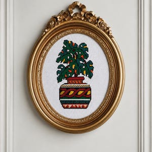 May include: A cross-stitch embroidery of a potted plant with dark green leaves and yellow accents, set in a decorative oval gold frame. The plant sits in a patterned pot featuring horizontal bands of red, yellow, green, and black geometric designs.