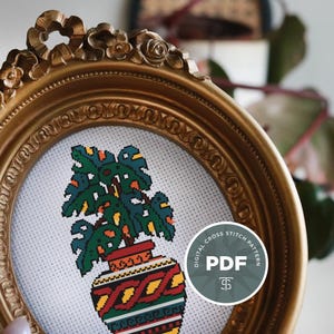 May include: A gold-coloured, ornate framed cross-stitch design of a potted plant with green leaves and a patterned pot. A circular sticker with the text "DIGITAL CROSS STITCH PATTERN PDF" is on the lower right. The frame has detailed floral embellishments.
